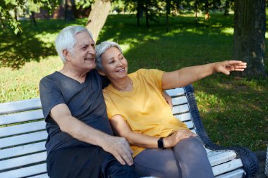 Pretty multi racial elderly 60s couple resting sit on bench in park after stroll, morning walk smiling enjoy talk relax outdoor discuss someone. Fit family, break, communication concept