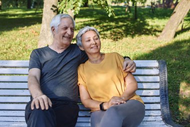 Mutlu çok ırklı yaşlı gri saçlı çift yürüyüş sonrası parkta bankta dinleniyor, gülerek gülümsüyor güneşli yaz gününde dışarıda dinleniyor. Uygun aile, kırık, aşk konsepti