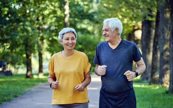 夏の公園で成熟した笑顔の白髪のフィットカップルジョギング 2人の年上のジョガーは 健康的なアクティブなライフスタイルを維持し 屋外で朝のアクティブな心臓をお楽しみください 体重減少 スポーツの概念 — ストック写真