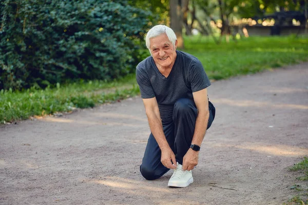 Elderly man in sportive wear ties shoelaces ready for morning jogging or sportive walk stroll in summer park smile look at camera. Healthy active lifestyle of retiree, work out