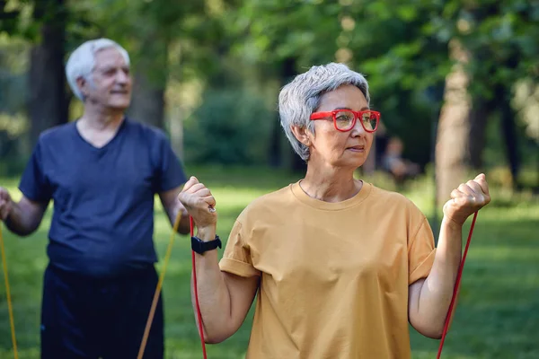年龄较大的60岁的夫妇穿着运动服进行双手练习 早上在夏季公园里用抵抗橡皮筋训练手臂 健康的生活方式 积极的退休生活使用现代器具 — 图库照片