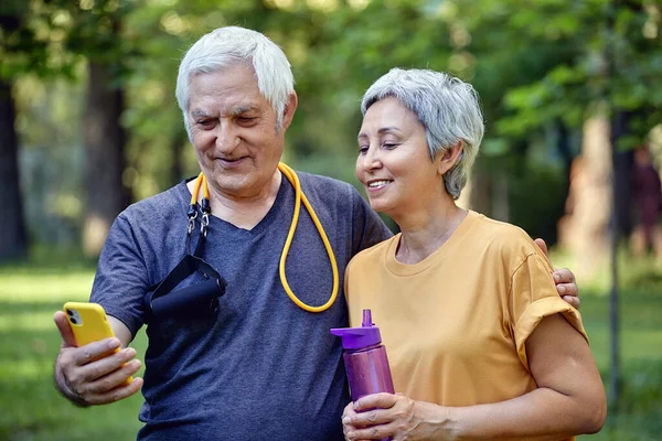 Old sportive couple staring at smart phone cam take selfie picture enjoy modern wireless tech usage while stroll in sportswear in summer park. Carefree retired life, lifestyle using technology concept