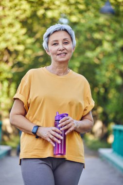 Spor kıyafetleri içinde yaşlı bir kadın, yaz parkında yaya yolunda duran ve sabah sporu yaparken susuzluğunu gideren tekrar kullanılabilir plastik şişeyi tutuyor. Sağlıklı yaşam tarzı