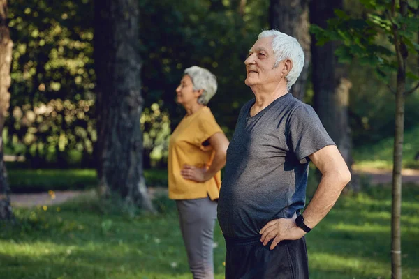 Senior spouses wear sportswear doing exercises outdoor in summer park in the morning. Healthy lifestyle, active retired life use modern appliance concept