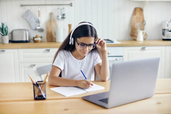 Pre-teen 12s girl in headphones and eyeglasses sit at table e-learns, listen online course, audio lesson, get new knowledge, skills using internet and modern tech. Development, education, eye-sight