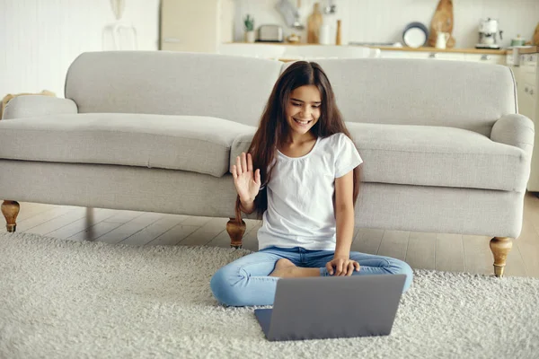 Attractive middle-eastern ethnicity pre-teen 12s girl sit on floor in living room wave hand greeting friend start videocall event use modern laptop. Virtual meeting, worldwide communication, blogging