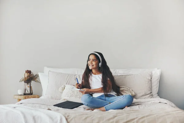 Beautiful pre-teen 12s girl in headphones listen audio course, e-studying seated on bed in bedroom alone at home. Homeschooling using modern tech, on-line class, modern wireless tech usage