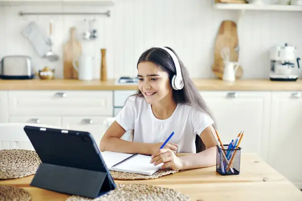 在无线耳机中的12岁以下的漂亮女孩坐在桌旁进行电子学习 听在线课程 听音频课 接受新知识 利用互联网和现代技术获得技能 儿童发展 图库图片