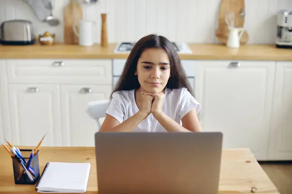 12岁以下的漂亮女孩坐在厨房的餐桌前 观看在线教育节目 新的录像或电影 独自呆在家里上网 年轻一代和现代技术在休闲中的应用 图库照片