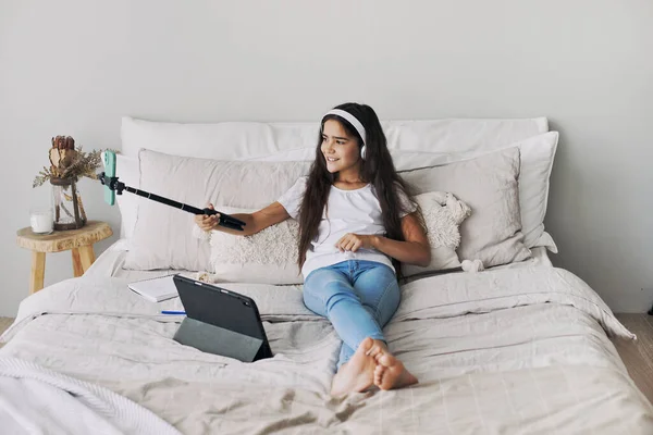 迷人的十多岁前女孩抱着自拍棒躺在床上拍照 为社交媒体频道录制视频 参加流媒体直播 在网上消闲 Vlogging Fun 免版税图库图片
