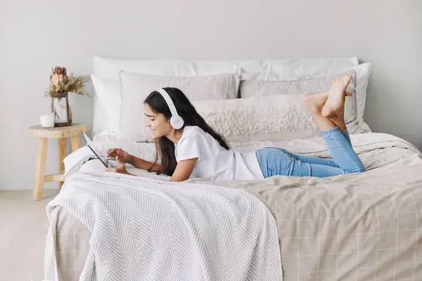 穿着无线耳机的可敬的十几岁前女孩躺在床上 床上躺着数字平板设备 年轻一代和现代技术的过度使用 周末在家里用小玩意娱乐 业余爱好和休闲 图库图片