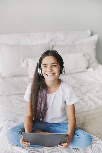 迷人的12岁前女孩穿着无线耳机 坐在舒适的卧室里 使用现代数码平板电脑 我为娱乐 教育和休闲而创造和使用技术 免版税图库照片
