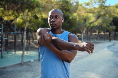 Siyah şortlu, kolsuz mavi gömlekli Afrikalı atletik adam parkta ısınma egzersizleri yapıyor, güneşli bir yaz günü. Spor, sağlıklı aktif yaşam tarzı, fiziksel eğitim konsepti