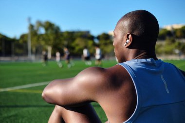 Genç sporcu adam kablosuz kulaklık takıyor, mavi atletik tişört giyiyor çimenli futbol sahasında oturuyor, maçtan sonra dinleniyor ya da antrenman yapıyor. Mola, spor, yaşam tarzı
