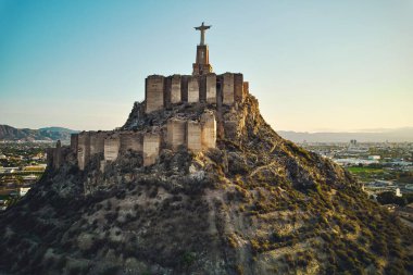 Engebeli tepenin tepesindeki Monteagudo heykeli, gün batımına bakan şehir manzarası. Antik kale ve çevreleyen manzara. Kültür ve tarihi sembol. Murcia, Alicante ili. İspanya
