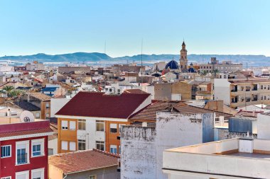 Novelda townscape with mountains view in background. Buildings rooftops, with clear blue skies and the mountainous terrain, typical Mediterranean architecture town, province of Alicante, Spain clipart