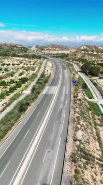 Parlak mavi gökyüzü ve dağınık bulutlar altında kuru, tepelik arazide kıvrılan çok şeritli bir otoyolun havadan görünüşü. Orihuela, İspanya, 