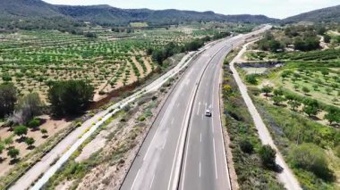 Parlak mavi gökyüzü ve dağınık bulutlar altında kuru, tepelik arazide kıvrılan çok şeritli bir otoyolun havadan görünüşü. Orihuela, İspanya, 