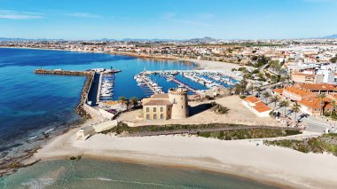 İspanyol Akdeniz kıyısındaki modern marina yakınlarındaki tarihi Torre de la Horada gözlem kulesinin havadan görünüşü kültürel miras, sahil mimarisi ve manzaralı kıyı güzelliğini birleştiriyor. İspanya