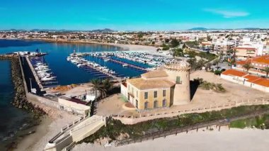 İspanyol Akdeniz kıyısındaki modern marina yakınlarındaki tarihi Torre de la Horada gözlem kulesinin havadan görünüşü kültürel miras, sahil mimarisi ve manzaralı kıyı güzelliğini birleştiriyor. İspanya