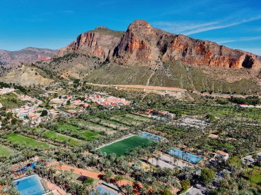 Palmeral de Orihuela 'nın ya da palmiye koruluğunun havadan görünüşü, kentsel yayılma ve berrak İspanyol gökyüzünün altındaki dağlık zemin altında yemyeşil bir vaha.