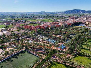 Palmeral de Orihuela 'nın ya da palmiye koruluğunun havadan görünüşü, kentsel yayılma ve berrak İspanyol gökyüzünün altındaki dağlık zemin altında yemyeşil bir vaha.
