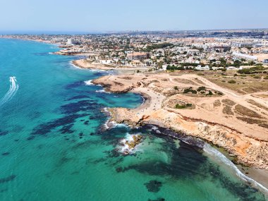 Kosta Blanca 'daki Playa Flamenca' nın havadan görünüşü, turkuaz Akdeniz suları, engebeli kayalık kıyı şeridi, kumlu koylar, yerleşim alanları ve canlı bir sahil tatil beldesi içeriyor.