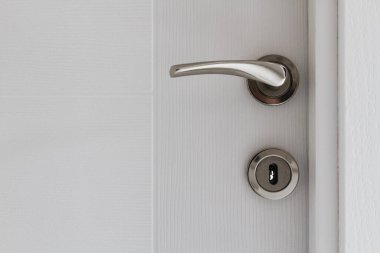 Silver door handle on an opened white door, revealing part of a grey wall. clipart