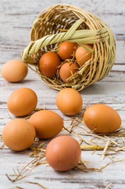 Taze kahverengi yumurtalar, biraz benekli, beyaz ahşap bir yüzeye saçılmış ve saman telleriyle çevrili, dokuma sepetinin ucunda.