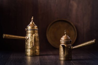 Two antique, ornate brass coffee pots placed on a dark wooden surface, pots having lids topped with a finial, round brass tray leaning against a dark wooden wall. clipart