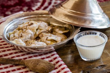 Geleneksel Çerçeve ravioli tabağı, bir bardak yoğurt, ahşap bir kaşık ve kırmızı beyaz çizgili bir bez eşliğinde metal bir kâsede servis edilir..