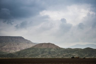 Bulutlu bir gökyüzünün altındaki tepelerin engin manzarası sessiz renklerle ve sakin bir atmosferle doğal arazinin huzurlu güzelliğini yakalıyor..