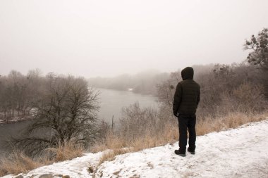 Siyah elbiseli dalgın bir adam kışın nehrin yanındaki yüksek bir uçurumda duruyor.