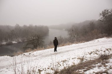 Siyah elbiseli dalgın bir adam kışın nehrin yanındaki yüksek bir uçurumda duruyor.