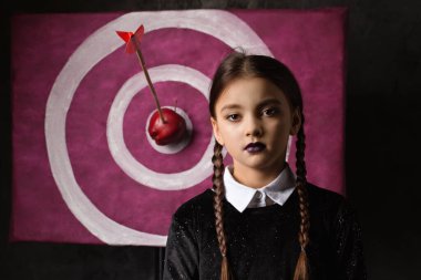 A gloomy girl with pigtails near a target with an arrow and an apple in a gothic style on a dark background
