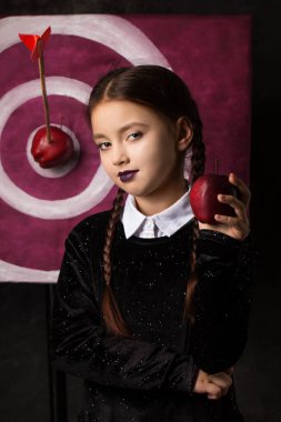 A gloomy girl with pigtails near a target with an arrow and an apple in a gothic style on a dark background