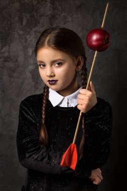 A gloomy girl with pigtails holds an arrow aimed at an apple on a dark background