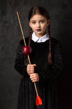 A gloomy girl with pigtails holds an arrow aimed at an apple on a dark background