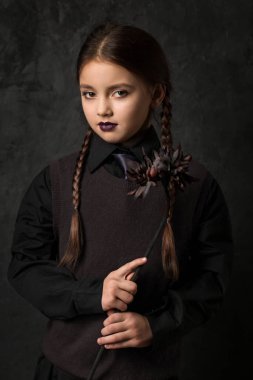 A sullen little girl with pigtails is holding a black dahlia flower. Gothic style on a dark background