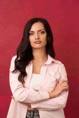 A beautiful, long-haired, self-confident brunette in a shirt folded her arms in front of her on a pink background