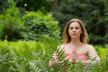 Genç ve güzel bir kadın yeşil ormandaki eğrelti otu yaprakları arasında meditasyon yapıyor.