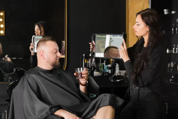 マスターの美容師は クライアントに鏡の中で新しい髪型を示しています 酒のグラスを持っている男 — ストック写真