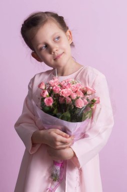 Pembe elbiseli şirin küçük bir kız pembe arka planda pembe güller tutuyor.
