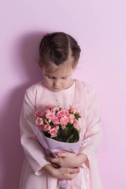 Pembe elbiseli şirin küçük bir kız pembe arka planda pembe güller tutuyor.