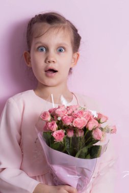 Pembe elbiseli şirin küçük bir kız pembe arka planda pembe güller tutuyor.