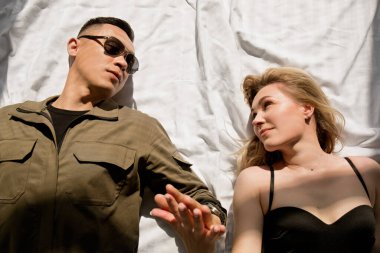 Üniformalı, güneş gözlüklü bir adam ve bir kadın güneşin altında bir yatakta el ele tutuşuyorlar.