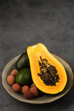 İki avokado, yarım papaya ve liçi üzümü gri arka planda elle yapılan gri bir tabakta.