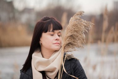 Bir kadın atkı takıyor ve elinde kuru bir miscanthus çiçeği tutuyor. Yüzünde bir gülümseme var.