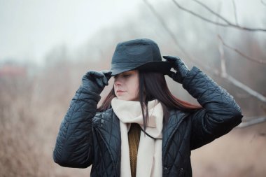 Siyah şapkalı ve siyah ceketli bir kadın ormanda duruyor. Eldiven ve eşarp takıyor.