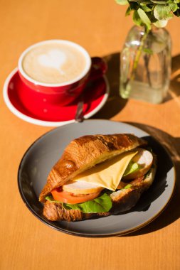 Jambonlu, marullu, domatesli ve peynirli bir sandviç kafedeki bir fincan kahvenin yanında duruyor.